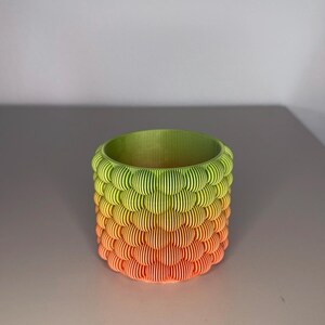 Peut inclure: Un pot de fleurs cylindrique imprimé en 3D avec un dégradé de couleurs. Le pot passe du vert en haut au jaune et à l'orange en bas. Le design présente un motif texturé en couches.
