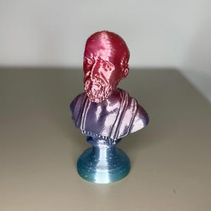 Peut inclure: Buste imprimé en 3D d'un homme barbu à l'expression sérieuse. Le buste présente un dégradé de couleurs, avec une tête et une barbe rouges, passant au violet et au bleu sur les épaules et la base. La sculpture est sur une surface claire.