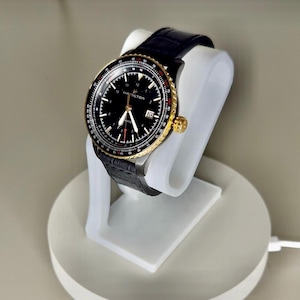 Peut inclure: Une montre Hamilton noire avec une lunette dorée et un bracelet en cuir noir est présentée sur un support de montre blanc. Le cadran de la montre est noir avec des marquages et des aiguilles blancs. C'est un article de luxe.