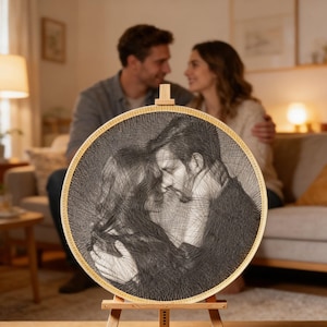 Puede incluir: Un retrato redondo de arte de hilo en blanco y negro de una pareja, exhibido en un caballete de madera. La obra de arte está enmarcada en un aro de madera natural. El fondo muestra a una pareja en un sofá.