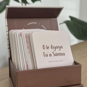 Può includere: Un mazzo di carte rettangolari intitolato O le Agaga Fa'a Sāmoa Samoan Value Cards, contenuto in una scatola aperta marrone. La carta superiore è beige chiaro con testo marrone scuro. L'interno del coperchio riporta le parole Kindness e Humility.