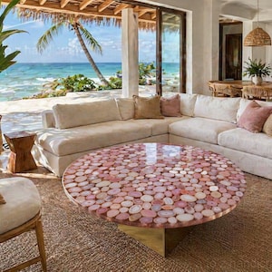 May include: A round coffee table with a mosaic top made of pink and white stones. The table sits on a gold base and is surrounded by a cream-colored sectional sofa and a woven rug. The room has a view of the ocean.