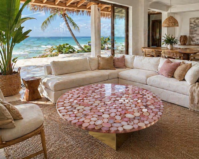 May include: A round coffee table with a mosaic top made of pink and white stones. The table sits on a gold base and is surrounded by a cream-colored sectional sofa and a woven rug. The room has a view of the ocean.
