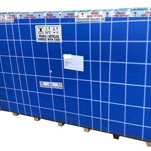 May include: Large, rectangular package wrapped in blue plastic with a grid pattern. White tape with "FRAGILE ARTICLES HANDLE WITH CARE" is visible. The package rests on wooden supports and has shipping labels.