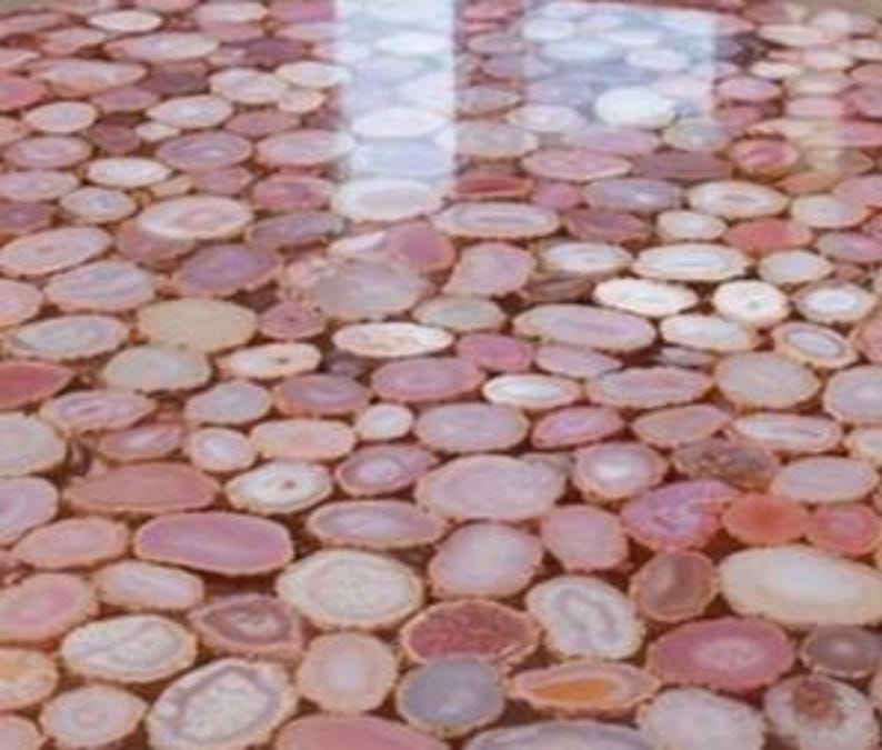 May include: A close-up view of a surface covered in polished, oval-shaped agate slices. The slices vary in shades of pink, beige, and white, creating a mosaic-like pattern. The surface reflects light, suggesting a glossy finish. The overall effect is decorative and natural.