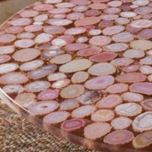 May include: A round table with a surface made of numerous, closely-packed, oval-shaped agate slices in shades of pink, peach, and cream. The table's edge is visible, showing a smooth, polished finish. The table's diameter is approximately 36 inches.