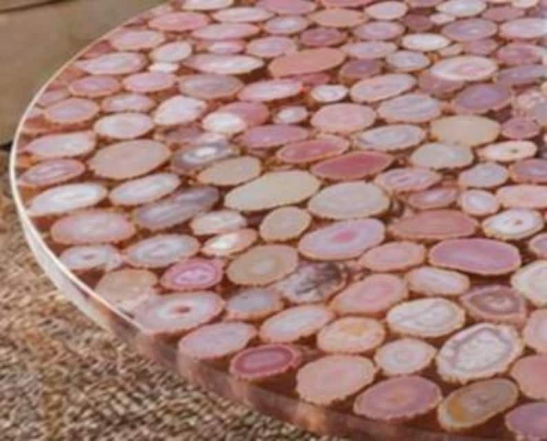 May include: A round table with a surface made of numerous, closely-packed, oval-shaped agate slices in shades of pink, peach, and cream. The table's edge is visible, showing a smooth, polished finish. The table's diameter is approximately 36 inches.