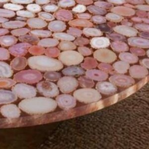 May include: Close-up of a round table surface inlaid with numerous polished agate slices in shades of pink, peach, and cream. The slices are set in a clear resin, creating a smooth, durable surface. The table's edge is visible, showing the resin and agate layers.