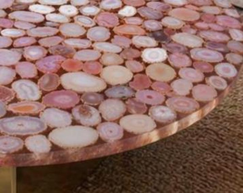 May include: Close-up of a round table surface inlaid with numerous polished agate slices in shades of pink, peach, and cream. The slices are set in a clear resin, creating a smooth, durable surface. The table's edge is visible, showing the resin and agate layers.