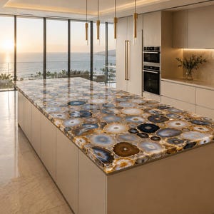 May include: A modern kitchen island with a countertop made of agate stone. The countertop features a pattern of blue, brown, and white circular designs. The island is beige and has a minimalist design. The kitchen has large windows with a view of the ocean.