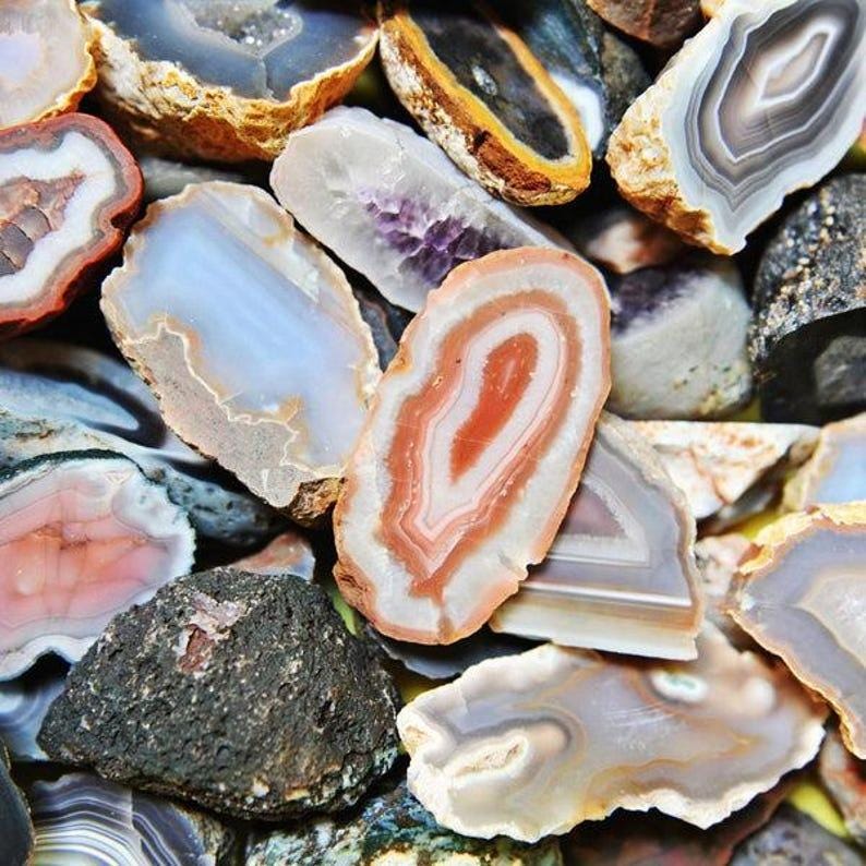 May include: A collection of polished agate stones in various colors and patterns. The stones display concentric bands of orange, white, gray, and blue. Some stones have a crystalline center, while others are solid.