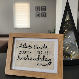 Puede incluir: Un cuadro bordado con las palabras "Alles Gute zum 10.!!! Hochzeitstag 10.10.25" en hilo negro sobre tela beige. El marco es de madera clara. La obra celebra un décimo aniversario de boda.