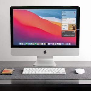 May include: A contemporary desk setup with an iMac computer displaying a vibrant screen. A white keyboard, mouse, and smartphone are on a grey felt mat. A grey mug and a small potted plant complete the workspace.