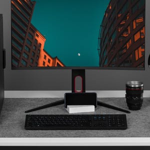 May include: A black computer setup featuring a monitor showing a cityscape with a teal sky. Includes a keyboard, mouse, headphones, and a coffee mug. The setup is on a white desk with a grey felt mat.