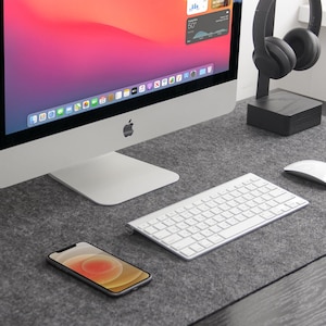 May include: A contemporary desk setup with a silver iMac computer, white keyboard, and wireless mouse. A smartphone and black headphones are also visible. The items are arranged on a grey felt desk mat.
