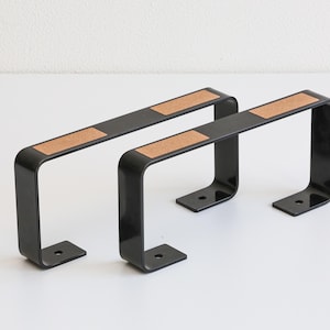 Peut inclure: Deux équerres d'étagère en métal noir avec un insert rectangulaire en liège. Les équerres ont une forme en U avec une base plate et une section supérieure pour le support de l'étagère. Les équerres sont conçues pour être montées sur un mur.