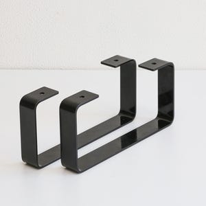 May include: Two black metal table legs in a U-shape design. Each leg has a flat top with pre-drilled holes for easy mounting. The legs are made of a sleek, durable metal and are suitable for various furniture projects.