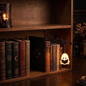 May include: A black metal bookend featuring an ornate scrollwork arm that extends outward, suspending a small, glowing white ghost figure holding a book by a chain. The ghost is illuminated from within, casting a soft light against the bookshelf.