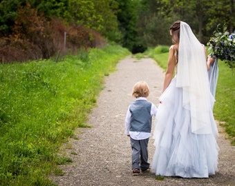 May include: A bride in a white dress and veil walks down a path with a young boy in a gray vest and pants. They are walking towards a bouquet of blue and green flowers.