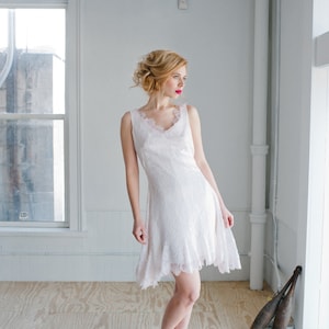 May include: A woman wearing a white lace dress with a V-neckline and a ruffled hem. She is standing in a room with white walls and a wooden floor. She is looking to the right and has red lipstick on.