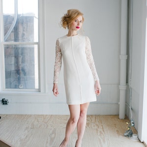 Op de afbeelding: Een witte jurk met kantmouwen. De jurk heeft een eenvoudige, aansluitende pasvorm en een ronde hals. Het model draagt witte schoenen.