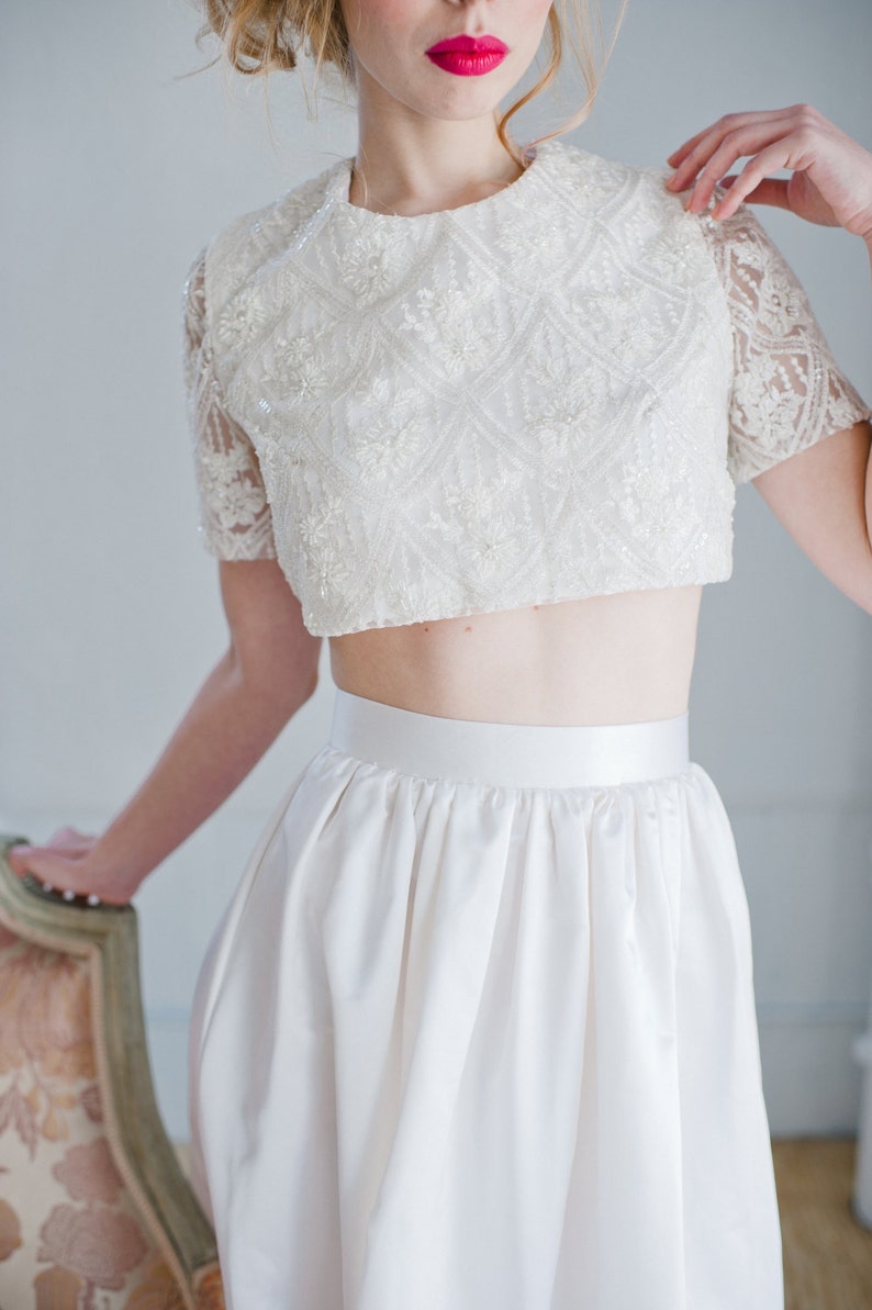 May include: A white crop top with a delicate lace design and a matching white skirt. The top has a round neckline and short sleeves.
