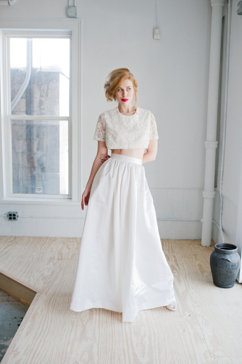 May include: A woman wearing a white lace crop top and a long white skirt. She is standing in a white room with a window in the background.