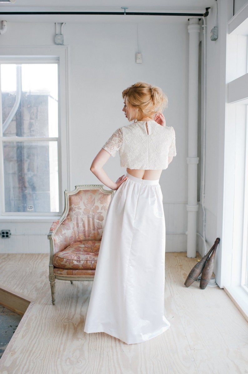 May include: A white lace crop top with a white skirt. The skirt is long and flowing, and the top has a fitted bodice and short sleeves.