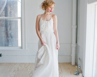 May include: A woman in a white sleeveless maxi dress with a ruffled neckline stands in a white room with a wood floor. She is looking down at the floor and her hair is pulled back.