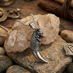 Pode incluir: Um pingente de metal prateado em forma de presa ou dente curvo, apresentando uma escultura intrincada de cabeça de lobo no topo com padrões detalhados de nós na lateral, suspenso por uma corrente de bolinhas prateada correspondente.