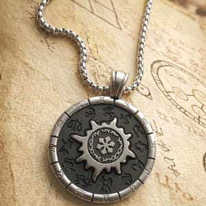 May include: Silver-tone pendant necklace featuring a circular pendant with a black center. The pendant has a sunburst design and zodiac symbols around the edge. The necklace is on a textured, tan surface.