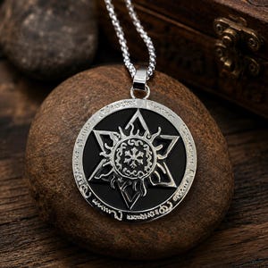 May include: A silver-toned circular pendant necklace featuring a black inlay with a six-pointed star design. The centre of the star displays a circular emblem with a small cross-like symbol. The outer silver rim is engraved with intricate text.