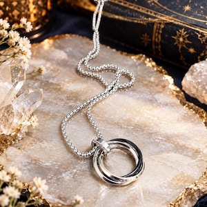 May include: A silver-toned necklace featuring a pendant composed of three interlocking rings. The pendant hangs from a round box chain. One of the rings is black, while the other two rings and the chain are polished silver-toned metal.