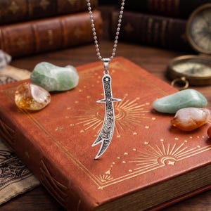 May include: A silver-toned pendant shaped like a curved sword or scimitar, featuring intricate black etched detailing on the blade and hilt. The pendant hangs from a silver-toned ball chain, showcasing a sharp, pointed tip and a cross-guard design.