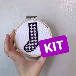 Puede incluir: Kit de punto de cruz con un diseño de calcetín con un patrón morado y blanco, presentado en un bastidor de bordado de madera. La palabra "KIT" está en una etiqueta morada. Fondo gris claro.