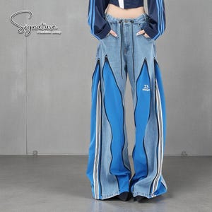 Puede incluir: Pantalones vaqueros azul claro con perneras anchas y un diseño único. Los pantalones presentan paneles azules vibrantes con rayas blancas y el número "23" impreso en una pierna. El logotipo "Signature Fashion Shop" es visible.