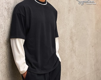 Puede incluir: Camiseta negra de manga corta sobre una camiseta crema de manga larga. El modelo lleva un collar de cadena plateada y pantalones negros. El fondo es una pared texturizada de tonos neutros. La imagen es de una tienda de moda.