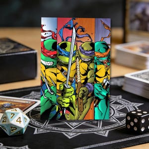 Puede incluir: Ilustración colorida de las Tortugas Ninja, cada una con su color de máscara característico: rojo, azul, morado y naranja. Sostienen sus armas. La imagen está sobre una superficie de madera con accesorios de juego.