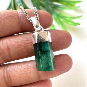 May include: A silver necklace featuring a raw, emerald green emerald pendant. The emerald is a rectangular shape with visible natural textures. The pendant is attached to a delicate silver chain, creating a simple and elegant piece of jewelry.