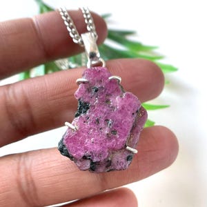 May include: A raw, pink ruby pendant necklace. The irregularly shaped gemstone is a vibrant magenta color with black inclusions. The pendant is attached to a silver chain. The necklace is held in a hand.