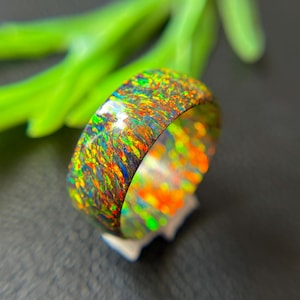 May include: A band ring featuring a dark base with a vibrant, multi-colored iridescent inlay. The surface displays a dense pattern of shimmering flecks in shades of bright green, fiery orange, and deep blue, creating a reflective, polished finish.