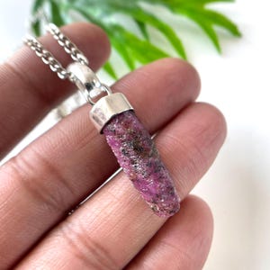 Peut inclure: Un collier avec pendentif en rubis brut. Le pendentif est une pierre pr&eacute;cieuse rugueuse, allong&eacute;e, rouge-ros&eacute; avec des inclusions sombres, sertie dans un capuchon en m&eacute;tal argent&eacute;. La cha&icirc;ne du collier est &eacute;galement de couleur argent&eacute;e. Le fond est flou avec du feuillage vert.