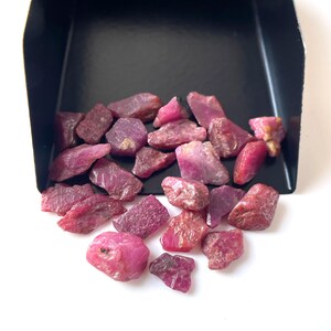 Può includere: Una collezione di pietre preziose di rubino grezze in varie tonalità di rosa e rosso, che fuoriescono da un vassoio nero. Le pietre grezze hanno un aspetto naturale e non lucidato, con trame e sfaccettature visibili.