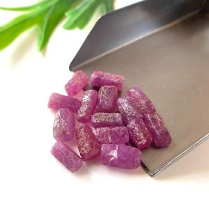 May include: A collection of rough, uncut ruby gemstones in varying shades of pink and red. The gemstones are displayed on a silver metal surface, with a few pieces scattered on a white background. Green leaves are visible in the background.