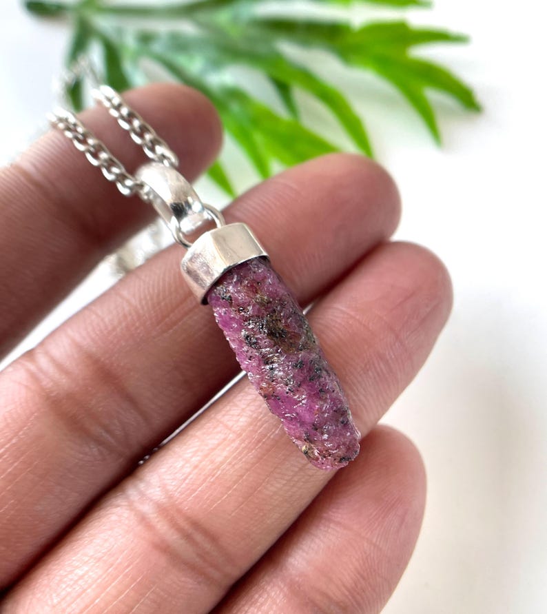 Peut inclure: Un collier avec pendentif en rubis brut. Le pendentif est une pierre pr&eacute;cieuse rugueuse, allong&eacute;e, rouge-ros&eacute; avec des inclusions sombres, sertie dans un capuchon en m&eacute;tal argent&eacute;. La cha&icirc;ne du collier est &eacute;galement de couleur argent&eacute;e. Le fond est flou avec du feuillage vert.