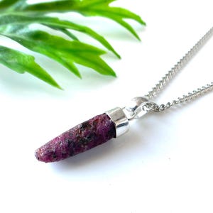 Peut inclure: Un collier en argent avec un pendentif en rubis brut. Le rubis est d'un rouge-rose profond avec des inclusions sombres. Le pendentif est attach&eacute; &agrave; une fine cha&icirc;ne en argent. Des feuilles vertes sont en arri&egrave;re-plan.