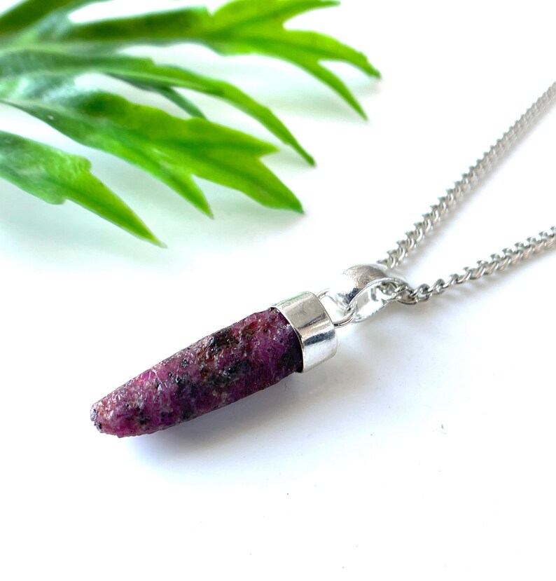 Peut inclure: Un collier en argent avec un pendentif en rubis brut. Le rubis est d'un rouge-rose profond avec des inclusions sombres. Le pendentif est attach&eacute; &agrave; une fine cha&icirc;ne en argent. Des feuilles vertes sont en arri&egrave;re-plan.