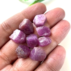 May include: A collection of eight small, raw, unpolished ruby gemstones with a deep magenta and pink hue. Each stone features an irregular, jagged shape with a textured, crystalline surface that reflects light, showcasing natural facets and variations in color intensity across the group.
