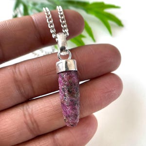 Peut inclure: Un collier pendentif en rubis brut. Le pendentif est un rubis cylindrique brut, dans des tons roses et rouges, avec des inclusions sombres. Il est attach&eacute; &agrave; une cha&icirc;ne de couleur argent&eacute;e. Le collier est tenu dans une main sur fond blanc.