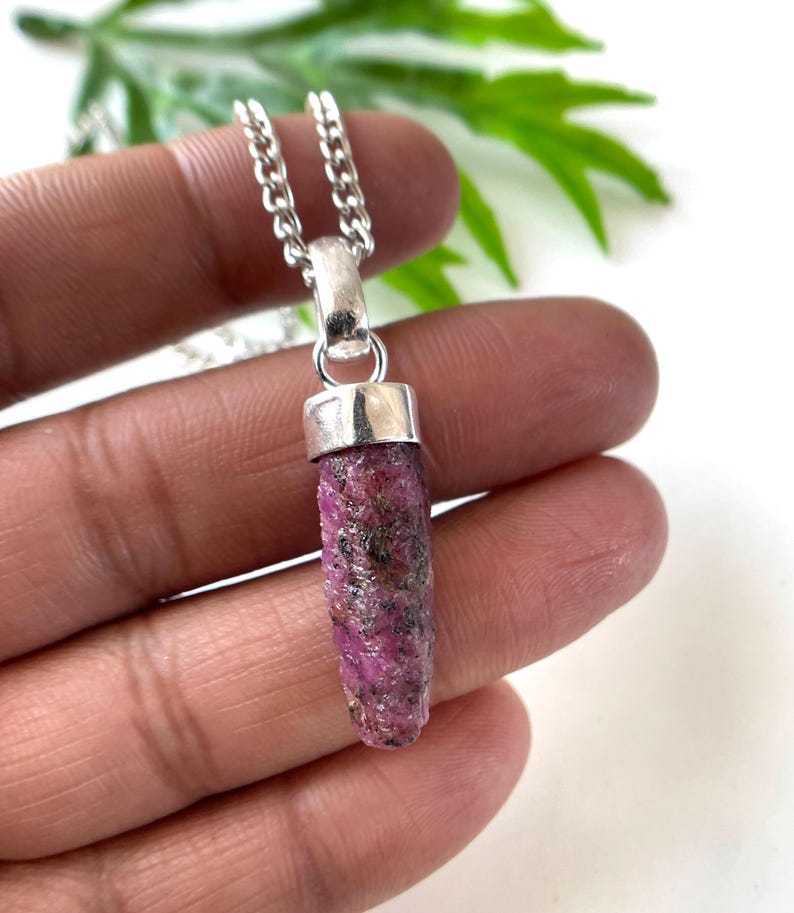 Peut inclure: Un collier pendentif en rubis brut. Le pendentif est un rubis cylindrique brut, dans des tons roses et rouges, avec des inclusions sombres. Il est attach&eacute; &agrave; une cha&icirc;ne de couleur argent&eacute;e. Le collier est tenu dans une main sur fond blanc.
